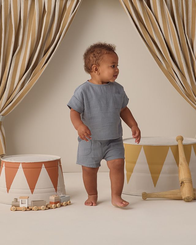 Baby in a blue outfit standing in a room with decorative curtains and toys.