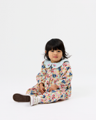 A toddler girl sitting on a white studio floor wearing a long-sleeve floral print jumpsuit with a light blue Peter Pan collar and white sneakers.