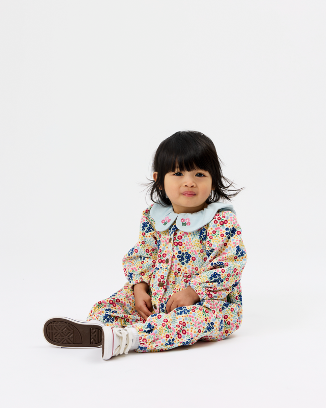 A toddler girl sitting on a white studio floor wearing a long-sleeve floral print jumpsuit with a light blue Peter Pan collar and white sneakers.