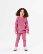 A young girl with curly brown hair stands against a plain white studio background wearing a matching two-piece pajama set with horizontal pink and light blue stripes, featuring a long-sleeve top and pants. She is wearing white sneakers.