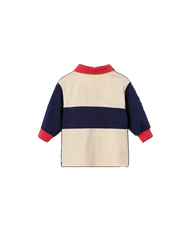 A long-sleeve colorblock rugby-style shirt shown from the back, featuring a cream body, a navy blue horizontal stripe, and red trim on the collar and cuffs.