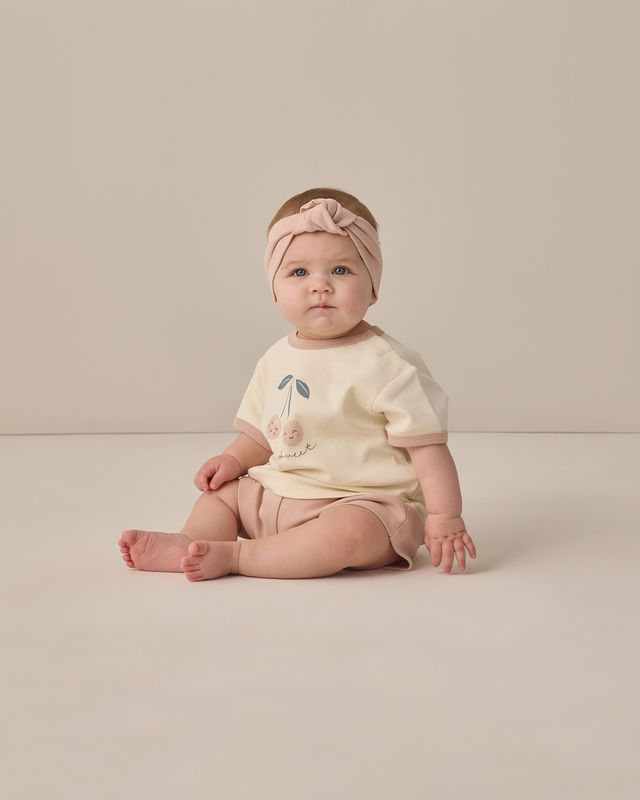 Baby sitting on a plain background wearing a light-colored outfit with a headband.