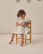 Child in a checkered dress sitting on a chair holding a toy, with a neutral background