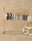 Wooden clothing rack with baby clothes against a beige wall, with decorative items on the floor.