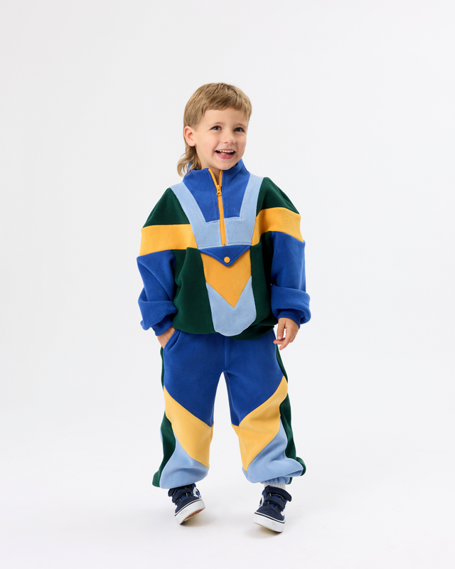 A young boy wearing a colorful, multi-panel fleece tracksuit with a half-zip pullover top and matching joggers, standing against a plain white studio background.