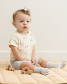 Baby sitting on a beige blanket with wooden toys, wearing a light-colored shirt and gray shorts.