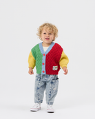 A toddler with curly blonde hair stands against a light gray studio background wearing a colorful cable-knit cardigan with green, red, yellow, and light blue sections, paired with light-wash jeans and white sneakers.