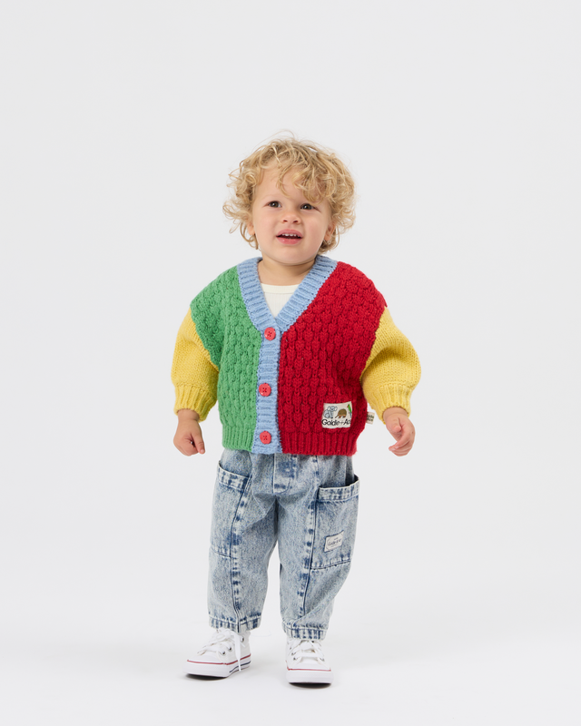 A toddler with curly blonde hair stands against a light gray studio background wearing a colorful cable-knit cardigan with green, red, yellow, and light blue sections, paired with light-wash jeans and white sneakers.