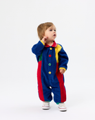 A toddler standing on a white studio background wearing a colorful patchwork corduroy one-piece jumpsuit with red, blue, yellow, and green panels, plus white sneakers.