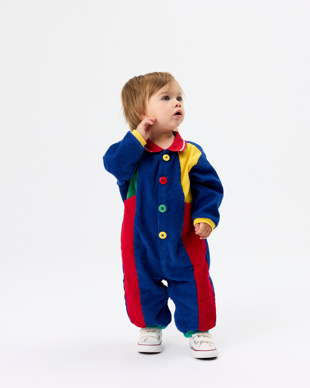 A toddler standing on a white studio background wearing a colorful patchwork corduroy one-piece jumpsuit with red, blue, yellow, and green panels, plus white sneakers.