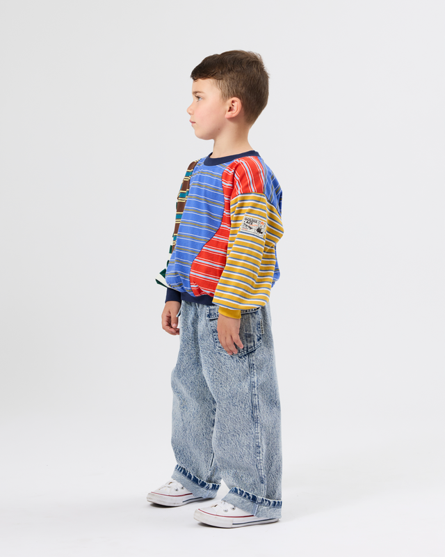 A young boy standing in profile wearing a colorful striped long-sleeve shirt, wide-leg light-wash jeans, and white sneakers against a plain light gray studio background.
