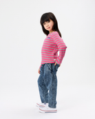 A young girl modeling a pink and blue striped long-sleeve knit top, paired with blue jeans and white sneakers, standing against a plain white studio background.