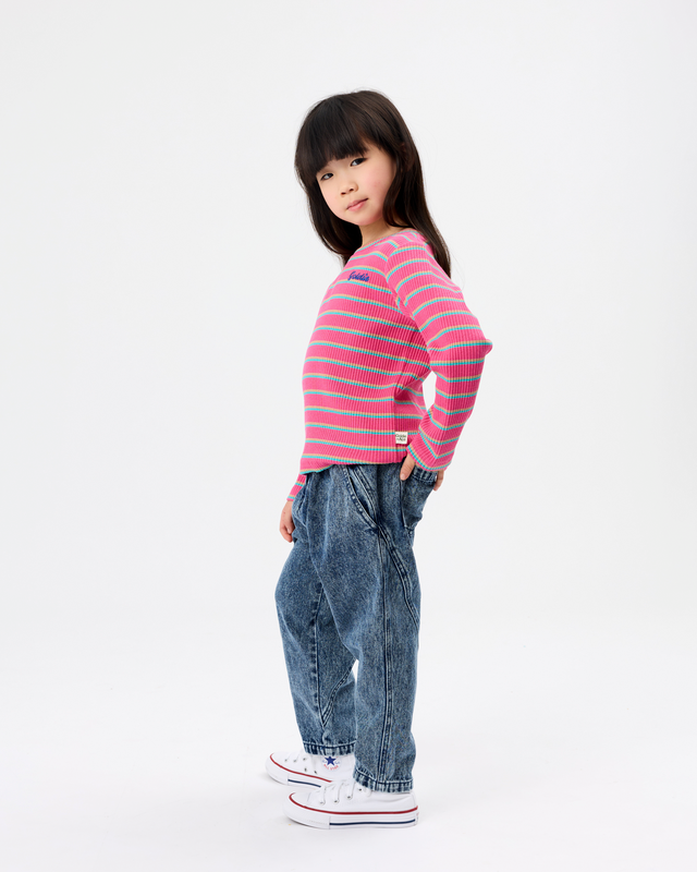 A young girl modeling a pink and blue striped long-sleeve knit top, paired with blue jeans and white sneakers, standing against a plain white studio background.