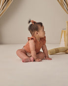 Baby in a rust-colored outfit sitting on a light surface with a neutral background