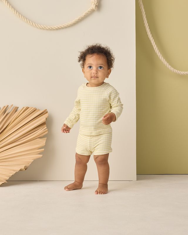 Baby in a cream-colored outfit standing in a minimalistic room with a wooden wall decoration.