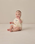 Baby sitting on a plain background wearing a light-colored outfit with a floral design.