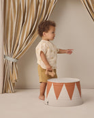 Child holding a teddy bear next to a decorative barrel with striped curtains in the background