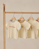 Three children's t-shirts on hangers against a beige wall.