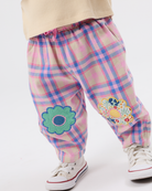 A close-up of a child's legs wearing pink and blue plaid pants with embroidered flower patches on the knees, paired with white sneakers and a cream ribbed top.
