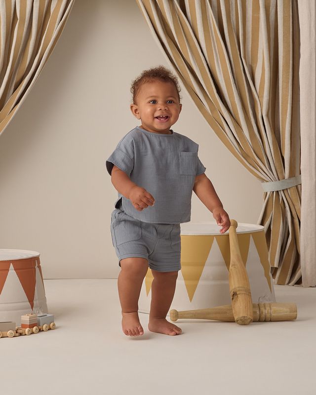 Child in gray outfit standing in a room with striped curtains and wooden toys.