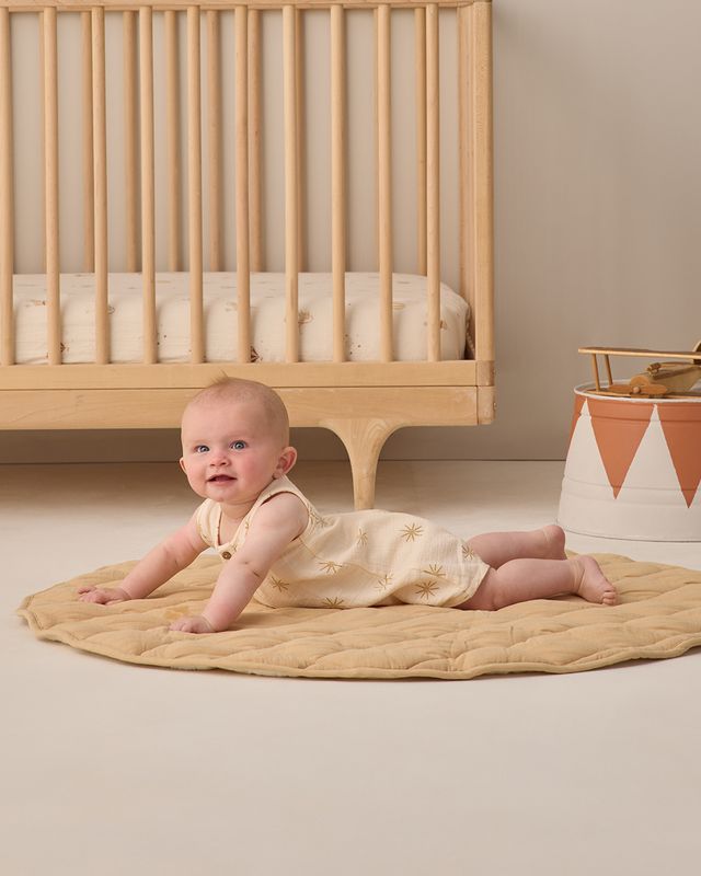 Baby crawling on a beige mat in front of a wooden crib with a neutral background