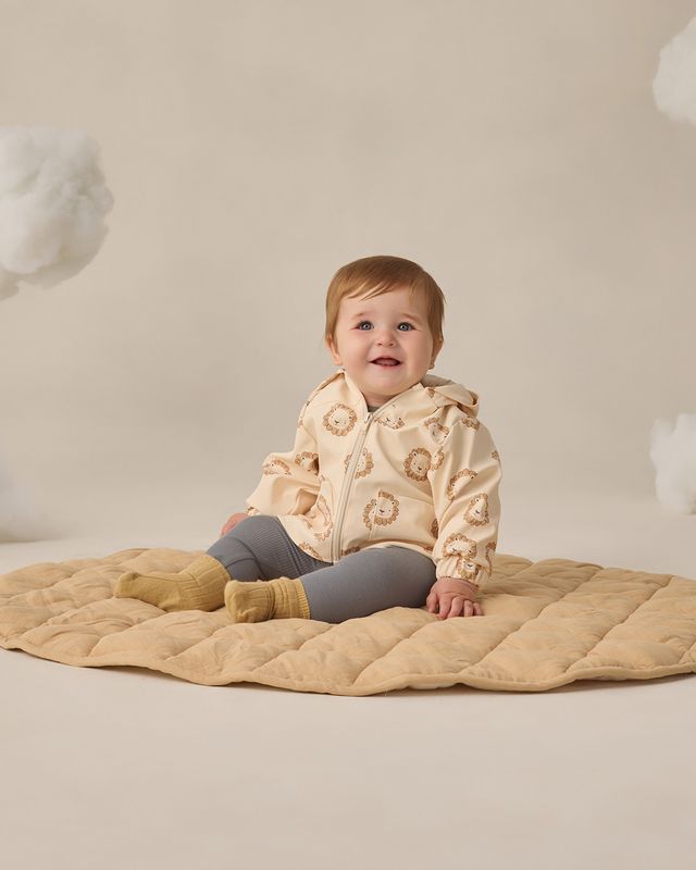Baby wearing a beige quilted jacket sitting on a beige blanket with a neutral background