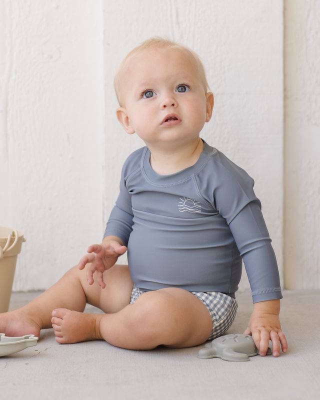 Baby wearing a blue long-sleeve shirt and checkered shorts sitting on a light-colored floor.