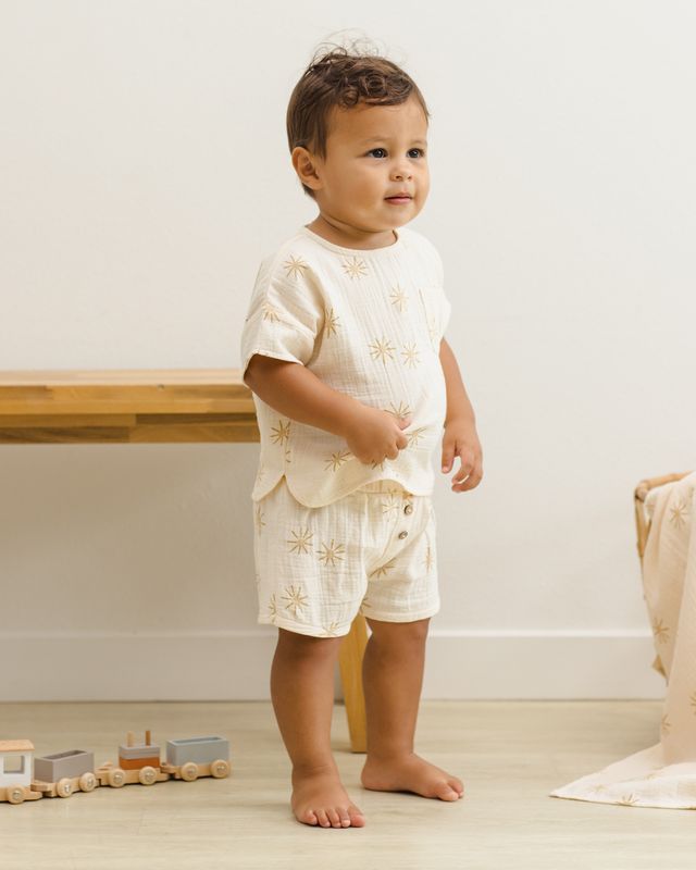 Baby in a white outfit with gold patterns standing on a light wooden floor.