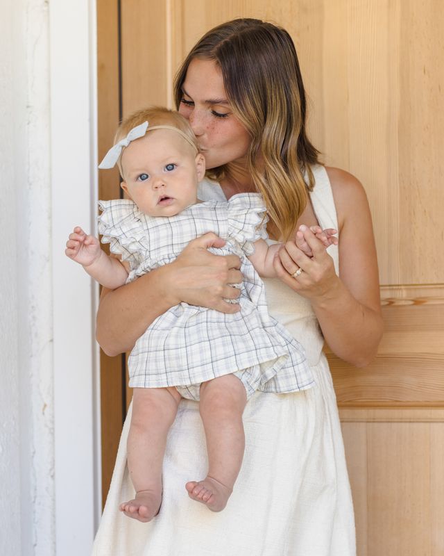 Woman holding a baby in a checkered outfit against a wooden background
