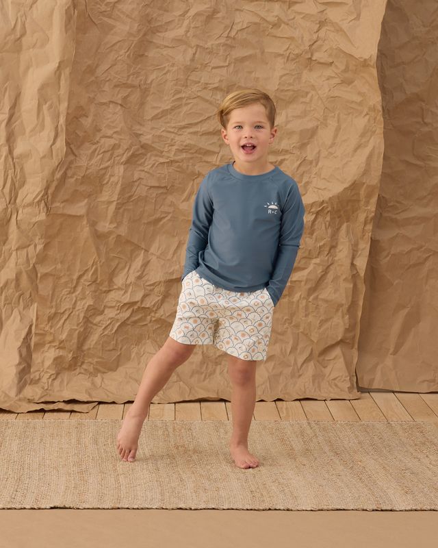 Child wearing a blue shirt and patterned shorts standing against a brown textured backdrop
