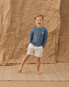 Child wearing a blue shirt and patterned shorts standing against a brown textured backdrop