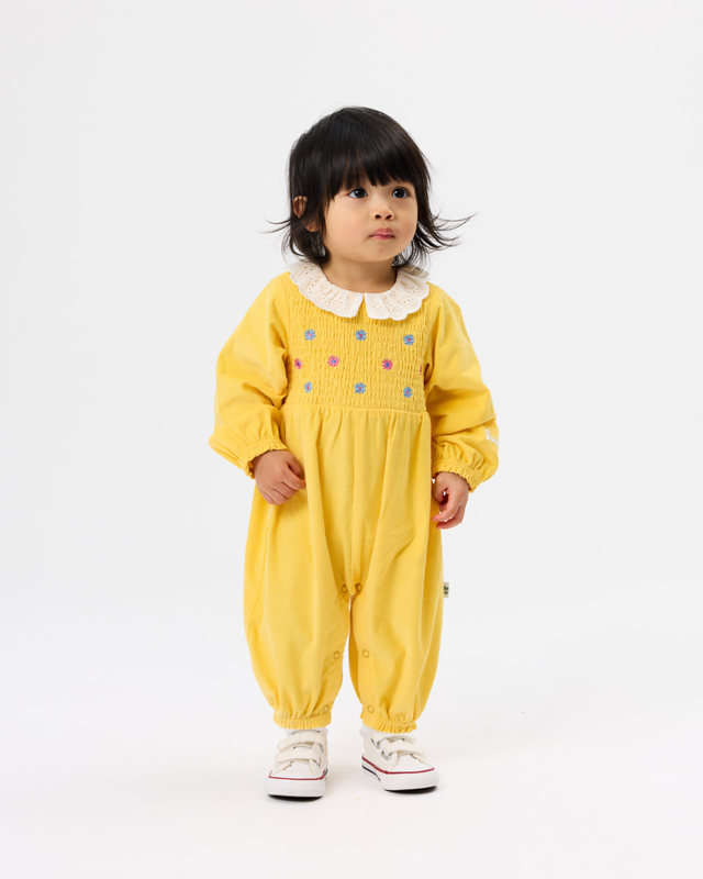 A toddler girl wearing a yellow smocked jumpsuit with a white ruffled Peter Pan collar and colorful embroidered dots, standing against a plain white studio background.