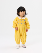A toddler girl wearing a yellow smocked jumpsuit with a white ruffled Peter Pan collar and colorful embroidered dots, standing against a plain white studio background.