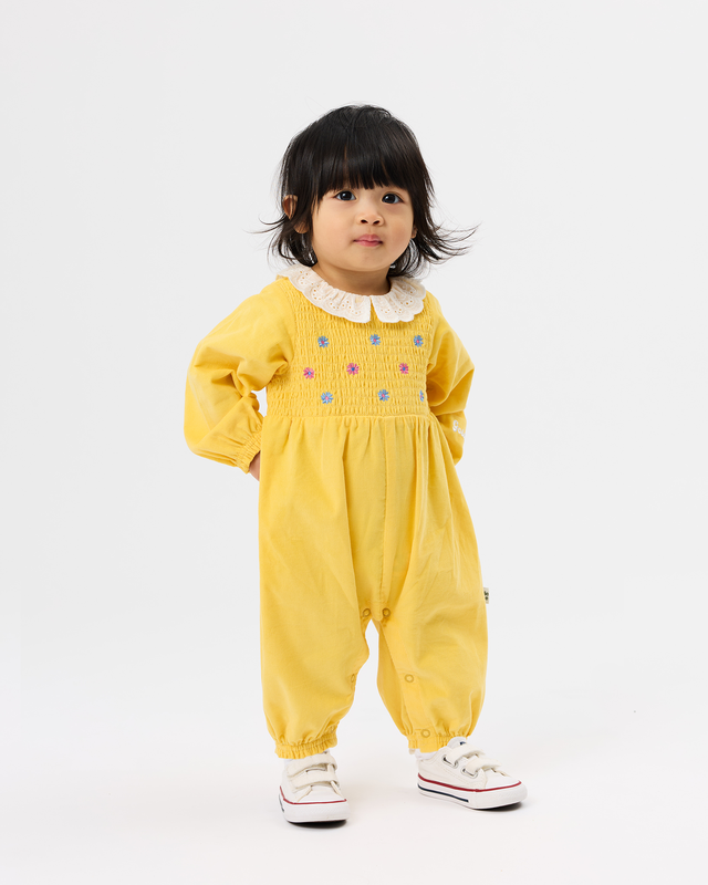 A toddler girl wearing a yellow smocked jumpsuit with a white ruffled collar and colorful embroidered dots, paired with white sneakers, standing against a plain white studio background.
