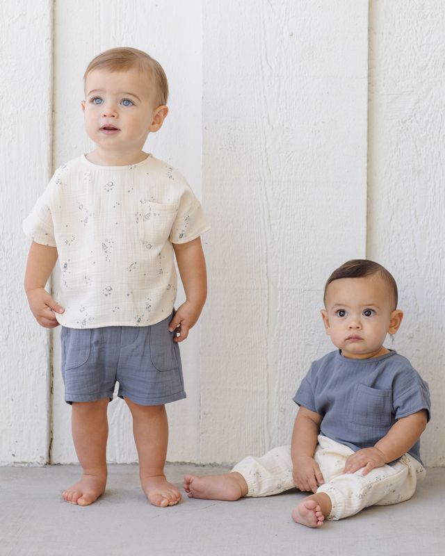 Two toddlers wearing light-colored clothing against a white wooden panel background