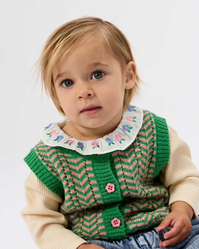 A toddler wearing a green and pink chevron knit vest over a cream ribbed long-sleeve top, featuring a white ruffled collar with colorful floral embroidery and pink buttons.