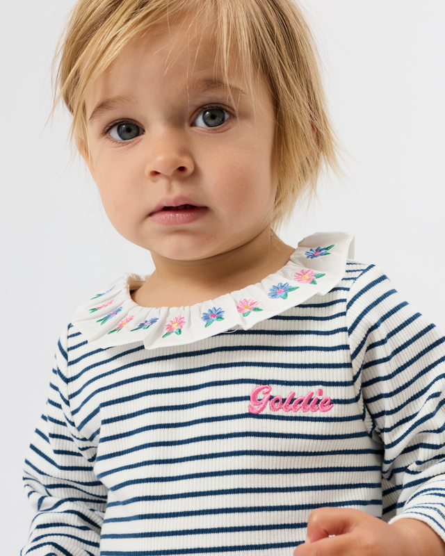 A close-up of a toddler wearing a navy and white striped long-sleeve top with a white ruffled collar decorated with small colorful flowers and a pink embroidered 'Grishie' logo on the chest, against a plain light gray background.