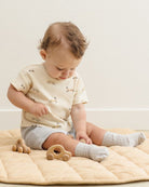 Baby sitting on a beige blanket with wooden toys, wearing a light-colored outfit.