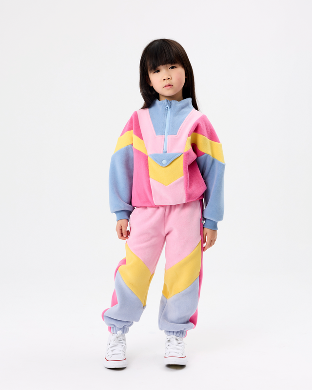 A young girl stands against a plain white studio background wearing a matching two-piece color-blocked tracksuit with pink, light blue, and yellow geometric panels, paired with white sneakers.