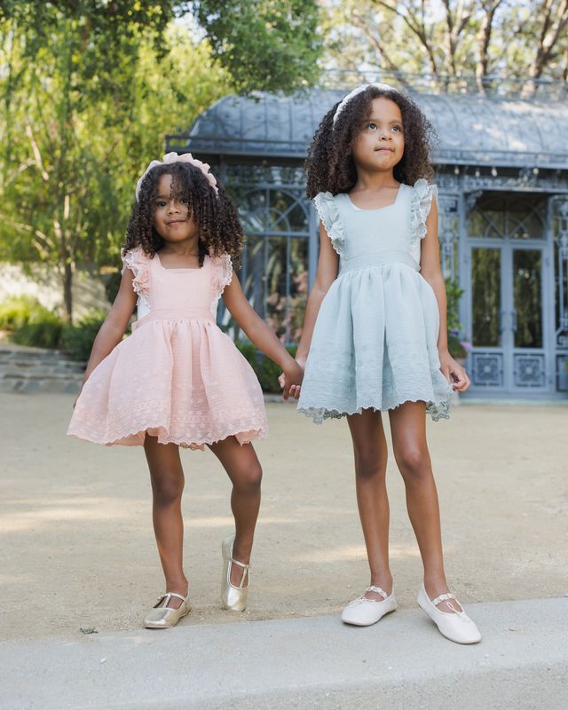 Two young girls in dresses standing outdoors with a building and trees in the background.