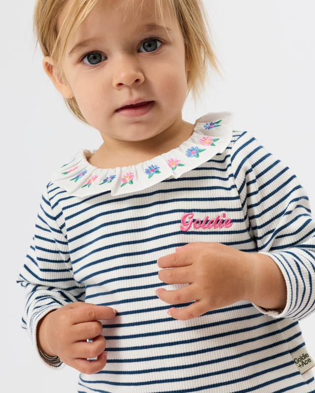 A close-up of a young child wearing a navy and white horizontally striped long-sleeve top with a white embroidered floral collar and a pink 'Gabbie' script on the chest.