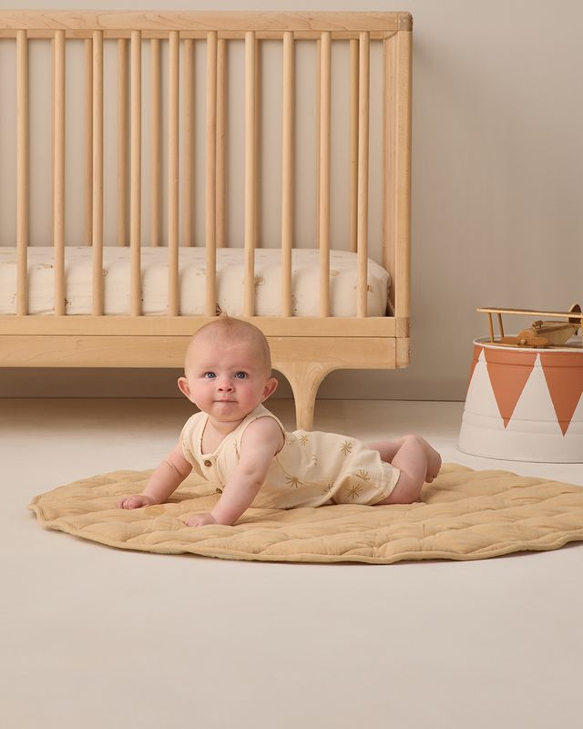 Baby lying on a beige blanket in front of a wooden crib in a nursery.