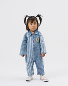 A toddler girl with dark hair in pigtails stands against a light gray studio background wearing a light blue denim one-piece jumpsuit with white vertical stripes, red buttons, and a circular patch on the chest, paired with white sneakers.
