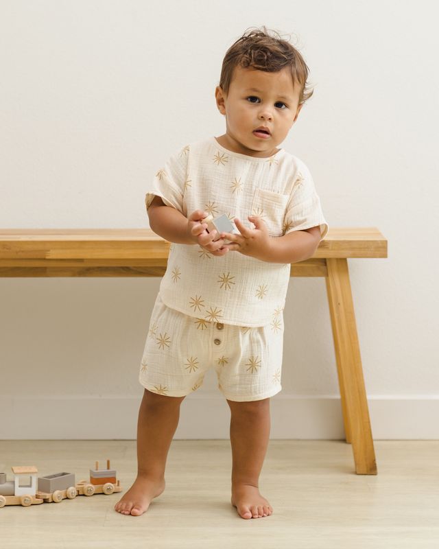Child wearing a cream-colored outfit with floral patterns standing in a minimalistic room.