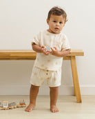 Child wearing a cream-colored outfit with floral patterns standing in a minimalistic room.