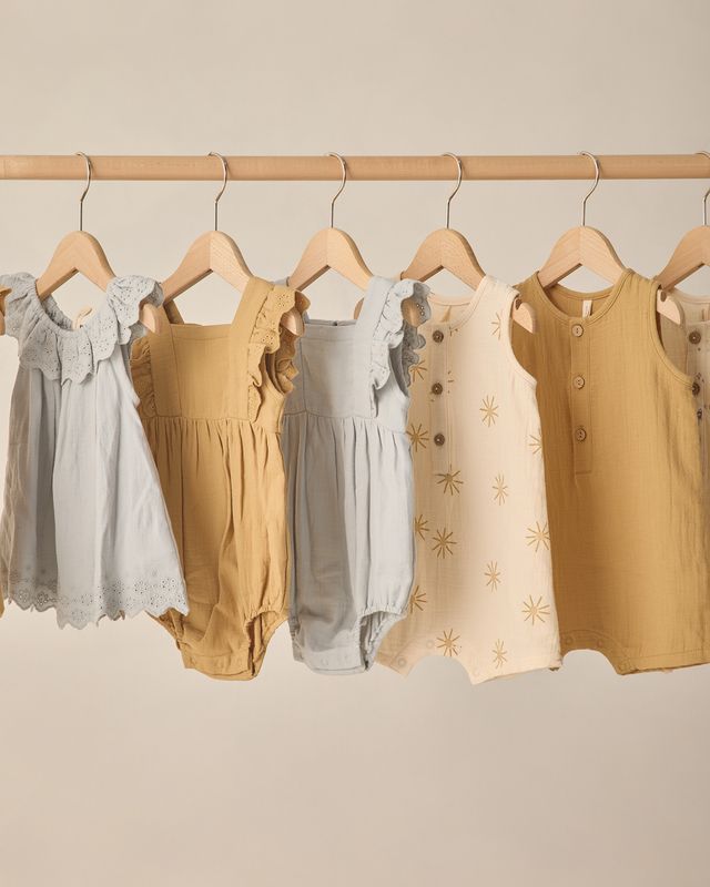Children's clothing including dresses and rompers on hangers against a neutral background