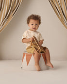 Child sitting on a small stool with a wooden toy, against a neutral background