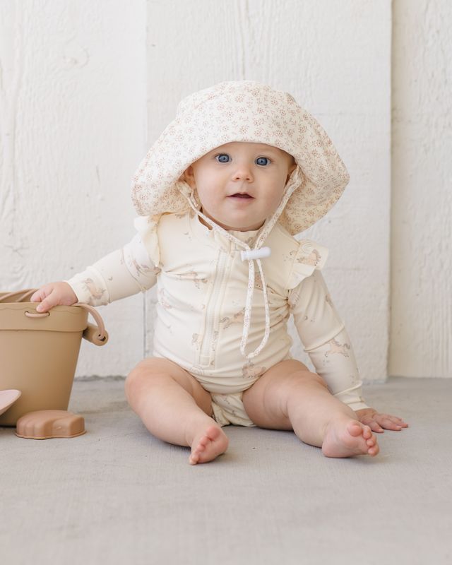 Baby in a cream-colored outfit with a hood sitting on a light surface.