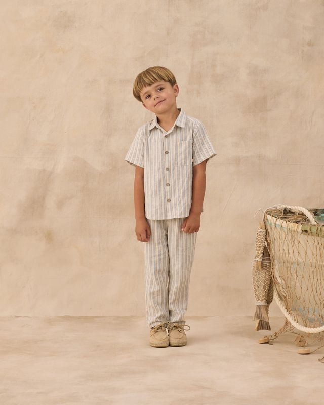 Young boy in striped shirt and pants standing next to a woven basket on a beige background