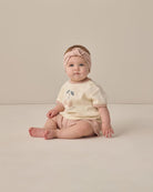 Baby sitting on a plain background wearing a light-colored outfit with a headband.